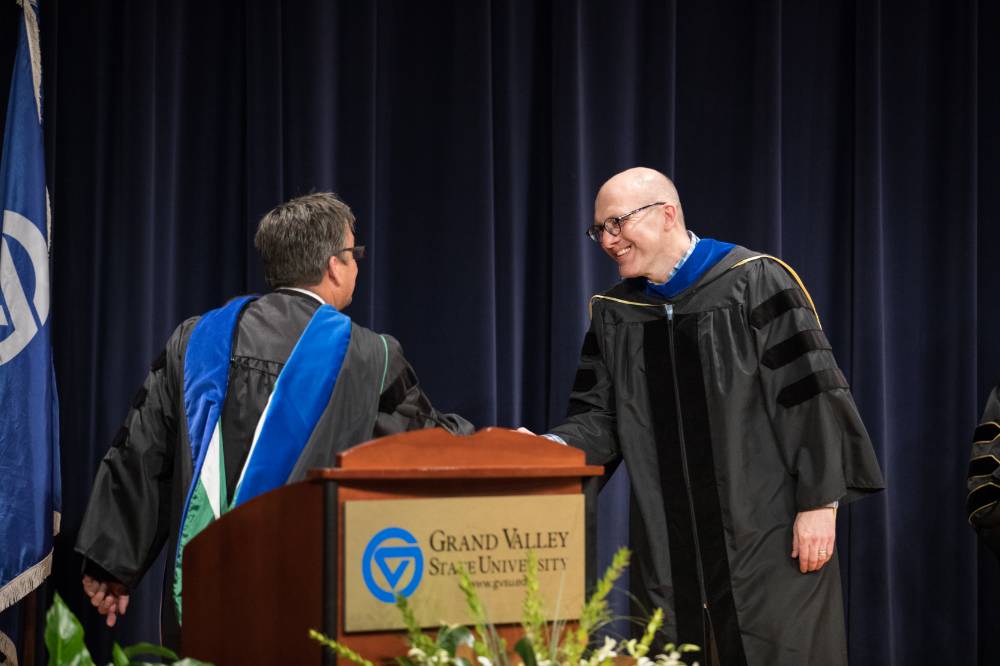 Two male faculty members share the stage and shake each others hands while smiling.